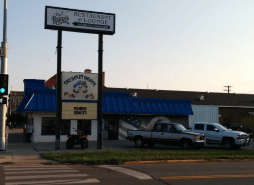 A building with a blue roof and sign for "The Donut Shoppe," featuring a pumpkin donuts advertisement. Two trucks parked outside.