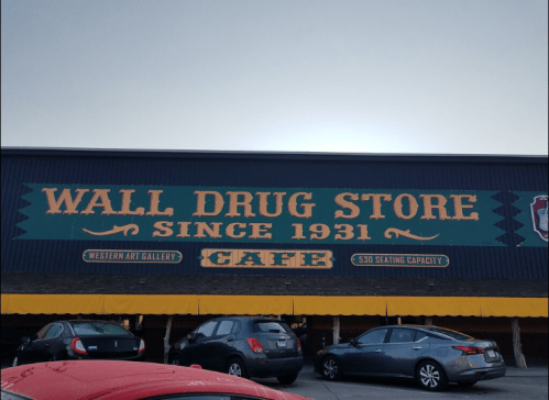 Sign for Wall Drug Store, established in 1931, featuring a café and western art gallery. Cars parked in front.
