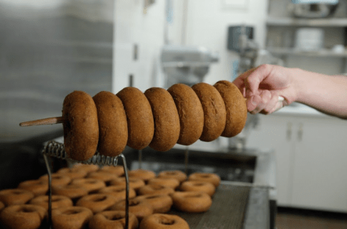 A hand holds a skewer of freshly made donuts above a tray of more donuts in a kitchen setting.