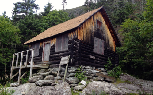 A rustic log cabin on a rocky foundation, surrounded by lush greenery and trees.