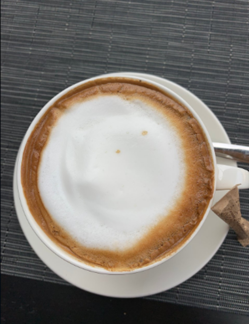 A close-up of a cup of coffee with frothy milk, resting on a white saucer against a textured black background.