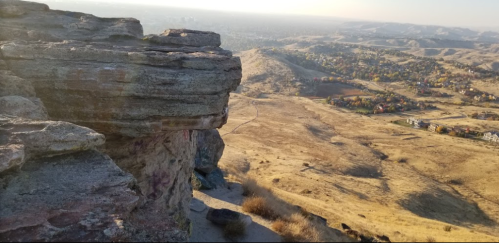 A rocky cliff overlooks a vast, golden landscape with rolling hills and a distant view of a town.