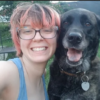A smiling person with pink hair poses with a happy black dog outdoors.