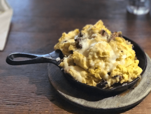 A cast iron skillet filled with cheesy scrambled eggs and chunks of meat, served on a wooden board.