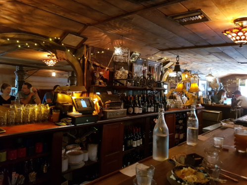 A cozy bar interior with wooden decor, shelves of wine, and patrons enjoying drinks in a warm, inviting atmosphere.