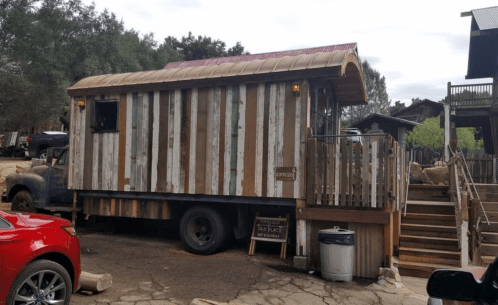 A rustic tiny house on wheels with a wooden exterior, parked beside a staircase and surrounded by trees.