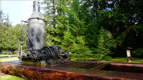Historic steam engine on a wooden platform, surrounded by lush green trees in a park setting.