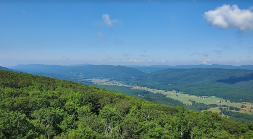 A panoramic view of rolling green mountains and valleys under a clear blue sky with scattered clouds.