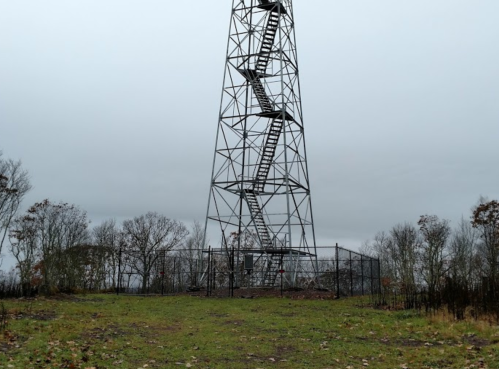 A tall metal tower with a spiral staircase, surrounded by a fence, set against a cloudy sky and sparse trees.