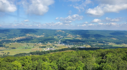 A panoramic view of rolling green hills and a small town under a blue sky with fluffy white clouds.