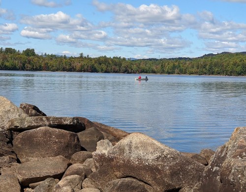 Two people in a kayak on a calm lake, surrounded by trees and rocky shoreline under a partly cloudy sky.