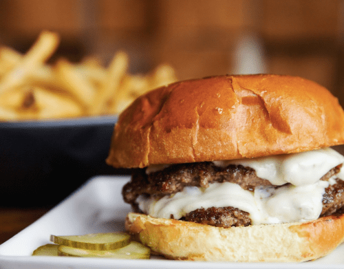 A juicy cheeseburger with melted cheese and pickles, served on a toasted bun, with a side of fries in the background.