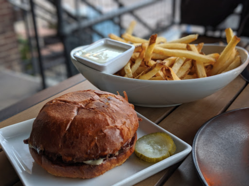 A delicious burger on a plate with a pickle, accompanied by a bowl of golden French fries and dipping sauce.