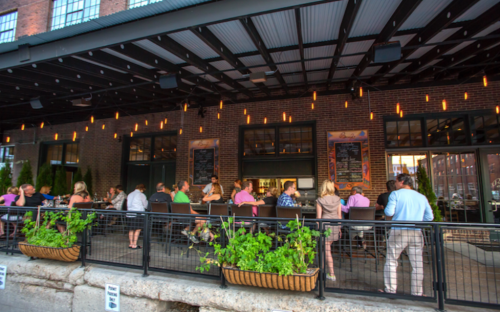 Outdoor dining area with people enjoying meals, surrounded by brick walls and hanging lights. Greenery in planters.