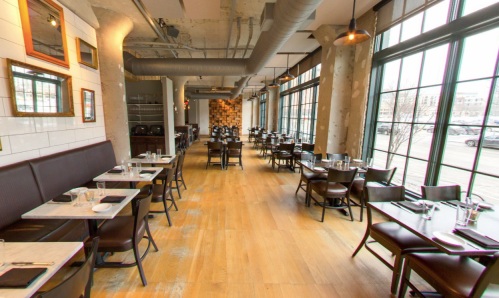 A spacious restaurant interior with wooden floors, large windows, and neatly arranged tables and chairs.