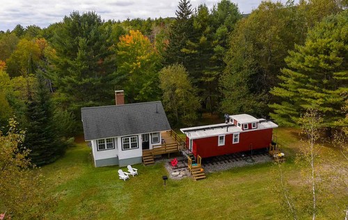 Aerial view of a cozy house and a red train car in a wooded area with green trees and a fire pit.