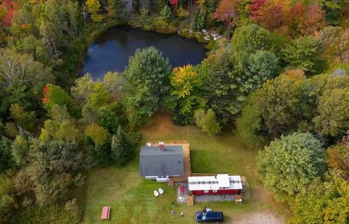 Aerial view of a house and trailer by a pond, surrounded by colorful autumn trees.
