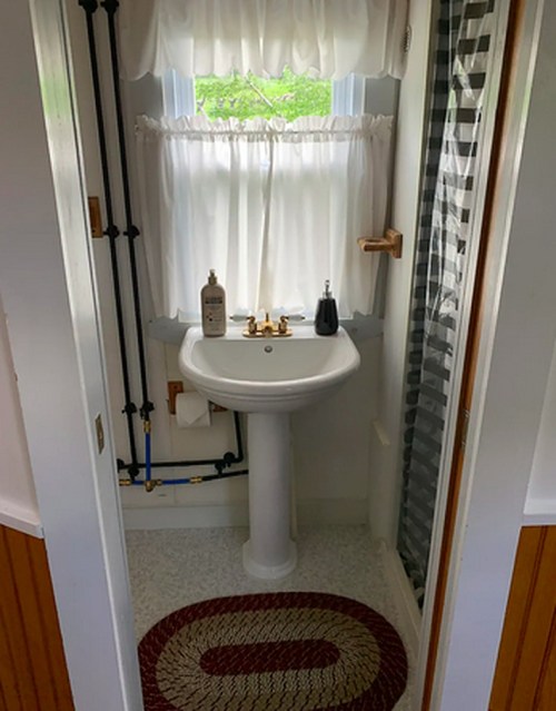 A small bathroom featuring a pedestal sink, shower, and a decorative rug on the floor. Natural light filters through a window.