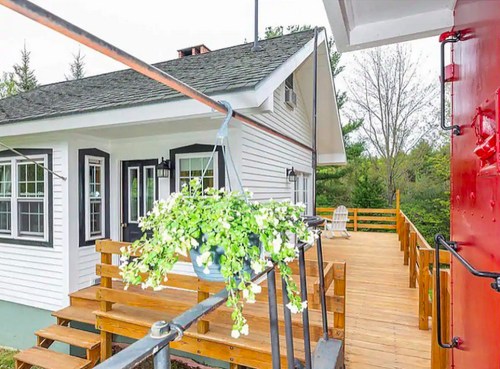 A charming house with a wooden deck, hanging plants, and a red structure nearby, surrounded by greenery.