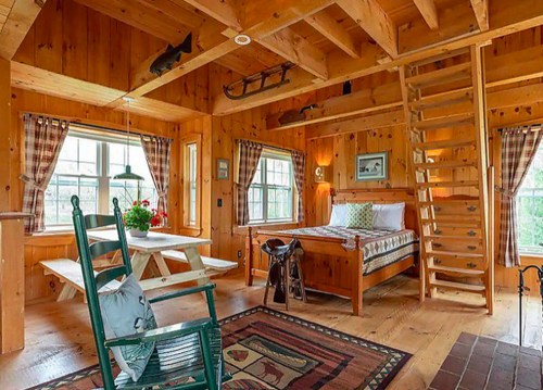 Cozy wooden cabin interior with a bed, dining table, rocking chair, and a staircase leading to a loft.
