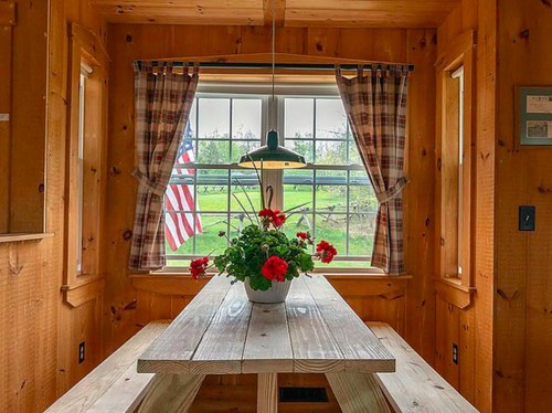Cozy wooden dining area with a table, potted red flowers, and a window featuring an American flag outside.