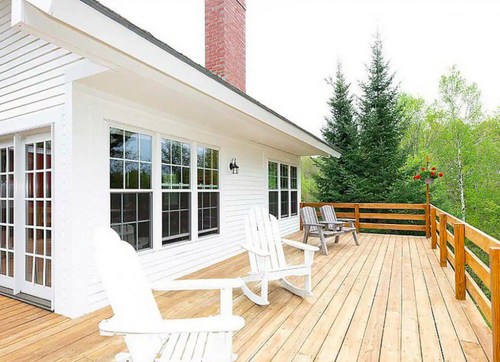A spacious wooden deck with white chairs, surrounded by greenery and a brick chimney on a house.