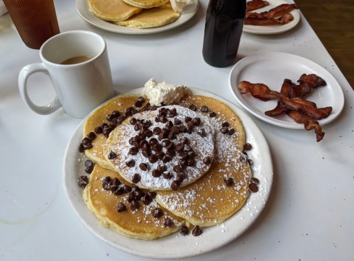 A plate of pancakes topped with whipped cream and chocolate chips, alongside a cup of coffee and a plate of bacon.