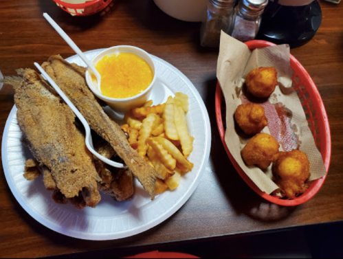 A plate with fried fish, crinkle-cut fries, and a cup of orange dessert, alongside a basket of hushpuppies.