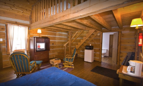 Cozy cabin interior with wooden walls, a loft, seating area, TV, and kitchenette. Warm lighting creates a welcoming atmosphere.