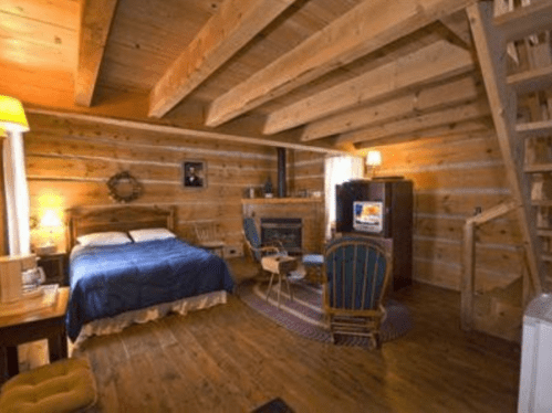 Cozy wooden cabin interior with a bed, seating area, TV, and staircase, featuring warm lighting and rustic decor.