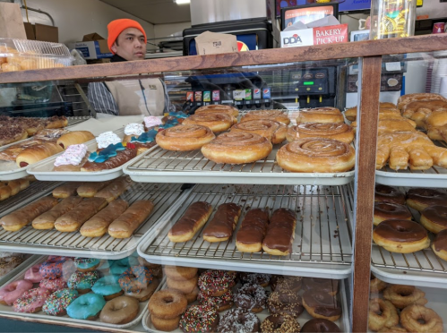 A bakery display case filled with various donuts and pastries, with a person in the background wearing an orange beanie.