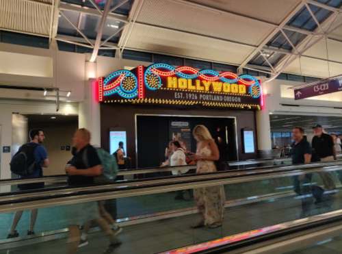 A bustling airport terminal with a colorful "Hollywood" sign, featuring travelers walking on a moving walkway.