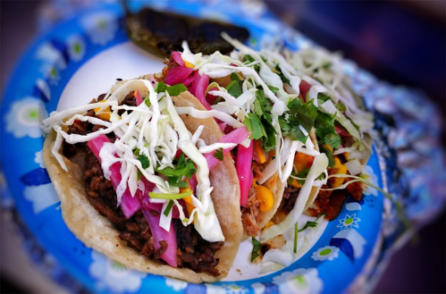 Two tacos on a paper plate, filled with meat, topped with cabbage, pickled onions, and cilantro, with a green pepper on the side.