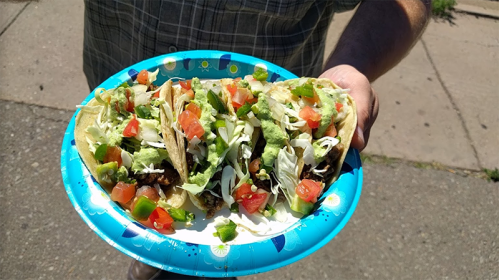 A person holding a plate with three tacos topped with lettuce, tomatoes, and green peppers on a colorful paper plate.