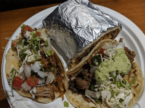 Three tacos on a plate: two with meat, onions, and toppings; one wrapped in foil.