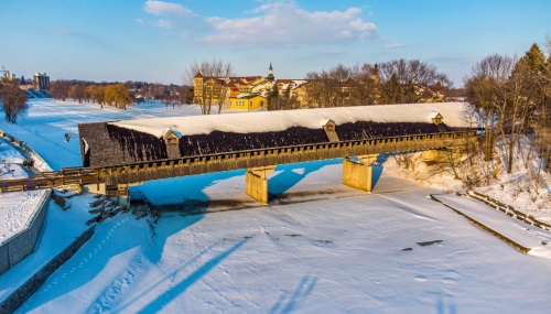 A snow-covered covered bridge spans a frozen river, with trees and buildings in the background under a clear blue sky.