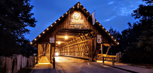 A lit wooden covered bridge at dusk, with a sign and road markings visible, surrounded by trees.