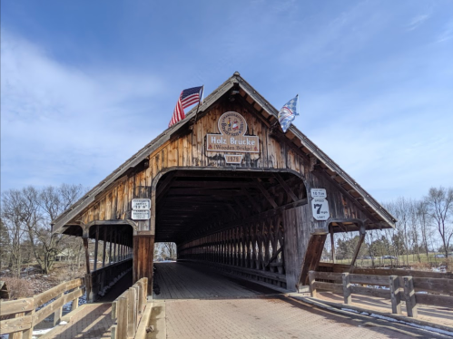 A wooden covered bridge with flags, featuring a sign that reads "Holz Brücke" and the year 1974.