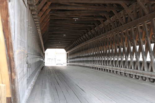 Interior view of a wooden covered bridge, showcasing its beams and wooden flooring, leading to a bright exit.