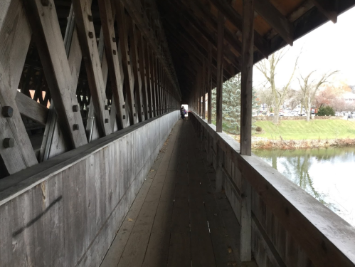 A long, wooden covered bridge with a view of a river and trees, leading to a bright opening at the far end.