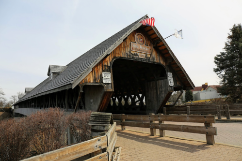 A wooden covered bridge with a peaked roof, featuring flags and signage, surrounded by a pathway and bare shrubs.