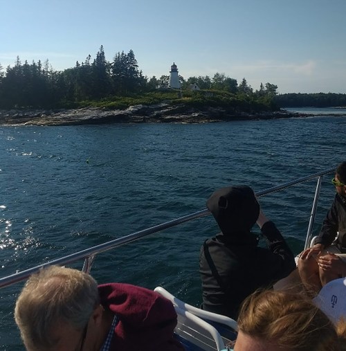 A boat passes by a rocky shoreline with a lighthouse surrounded by trees on a sunny day.
