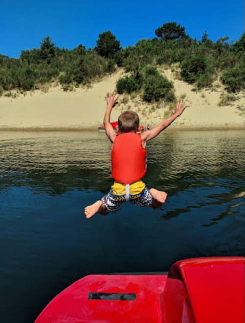 A child in a life jacket jumps off a boat into calm water, surrounded by trees and sandy hills.