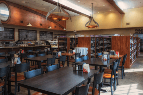 A spacious café with wooden tables, chairs, and shelves filled with books, featuring warm lighting and a welcoming atmosphere.