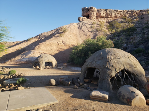 Two dome-shaped structures made of natural materials, set against a rocky hillside under a clear blue sky.