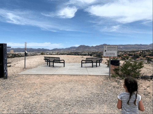 A child stands near a viewpoint with benches, overlooking a desert landscape and distant mountains under a blue sky.