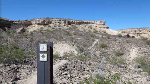 Trail marker in a rocky desert landscape with a clear blue sky and rugged terrain in the background.
