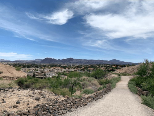 A scenic view of a desert landscape with a path leading to a valley of homes and mountains under a blue sky.
