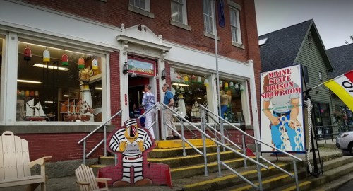 Two people stand outside a colorful store with a sign reading "Maine State Prison Showroom" and decorative items visible.