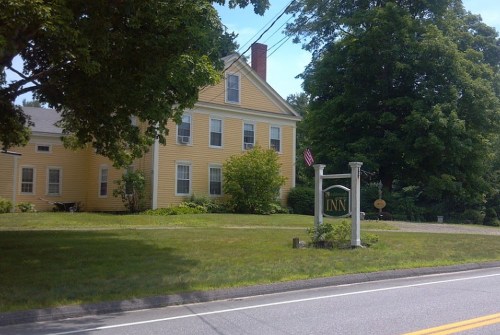 A yellow inn with a sign, surrounded by greenery and trees, located by a road with an American flag visible.